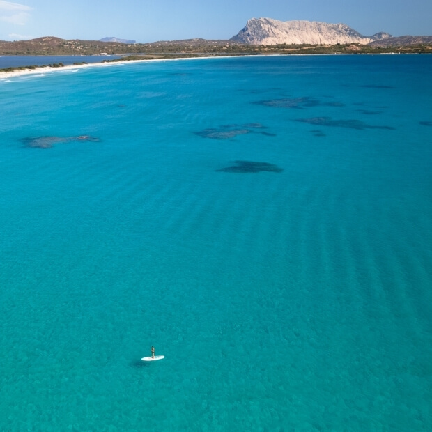 Immagine aerea di un paddleboarder su acque blu, con spiaggia bianca e colline verdi sullo sfondo sotto un cielo sereno.