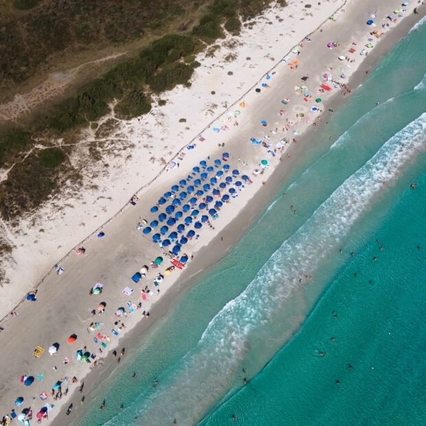 Vista aerea di una spiaggia affollata con persone sotto ombrelloni blu, l'oceano a destra e sentieri erbosi sullo sfondo.