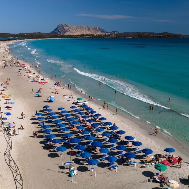 Scena di spiaggia vibrante con ombrelloni blu, lettini, acqua cristallina e paesaggi verdi sullo sfondo.