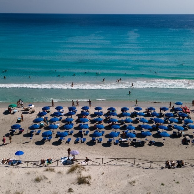 Una spiaggia serena con ombrelloni blu, persone che camminano, onde turchesi e surfisti in lontananza sotto un cielo blu.