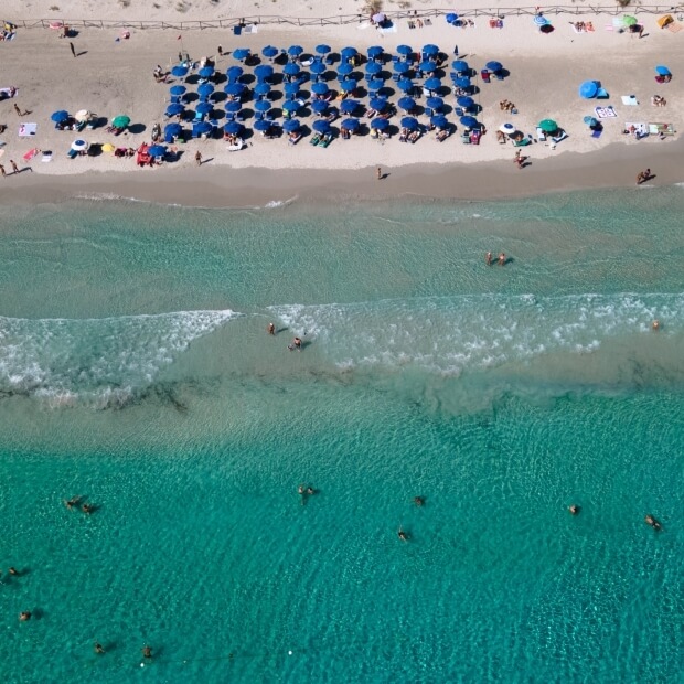 Vista aerea di una spiaggia affollata con ombrelloni blu, persone che si rilassano e nuotano in acque turchesi.