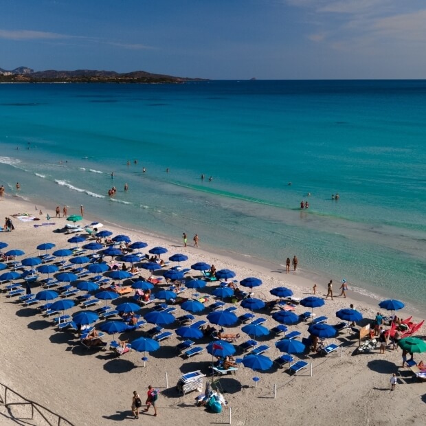 Spiaggia serena con ombrelloni blu, bagnanti e montagne sullo sfondo, cielo azzurro e atmosfera di relax.