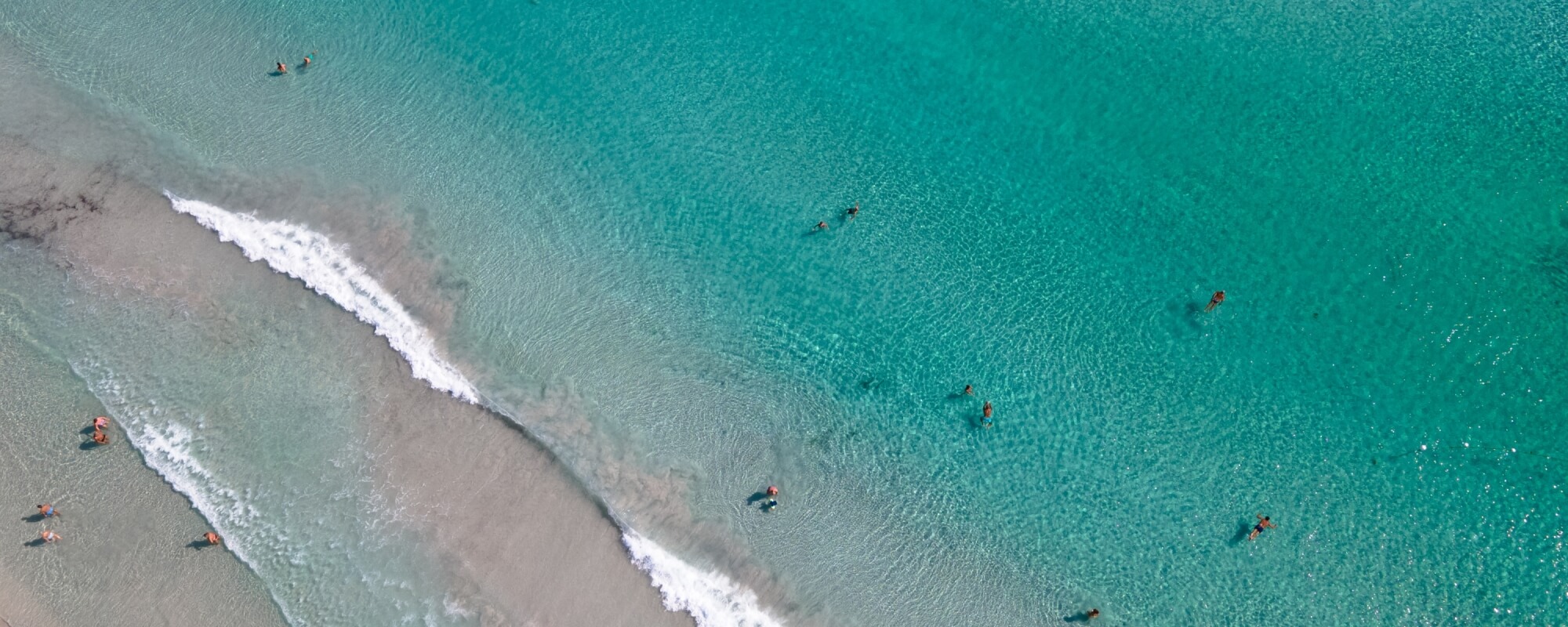 Spiaggia serena con persone che nuotano in acqua turchese, onde leggere e sabbia chiara lungo la riva.