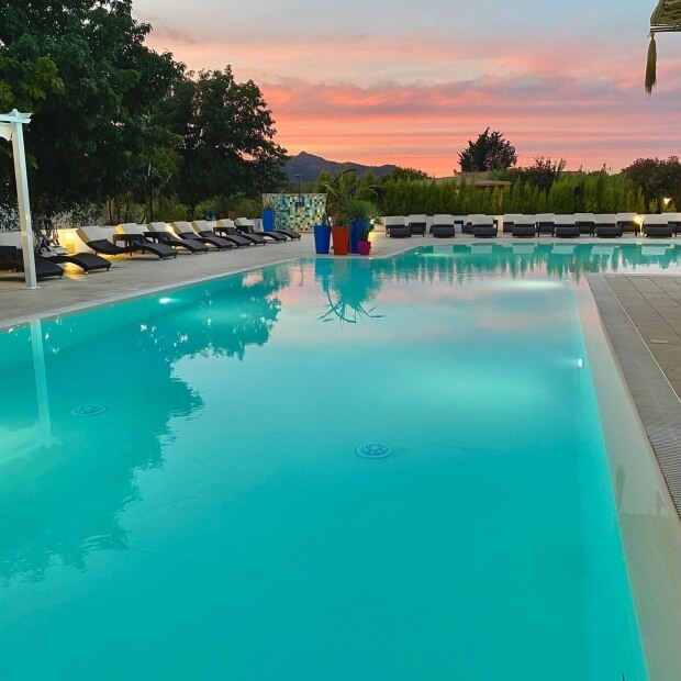 Una scena serena di un'area piscina al tramonto, con acqua blu, sedie a sdraio colorate e un cielo dipinto di arancione e viola.