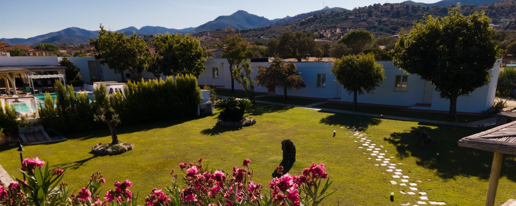 Una villa bianca circondata da un paesaggio verde, con piscina, fiori rosa e montagne sullo sfondo.