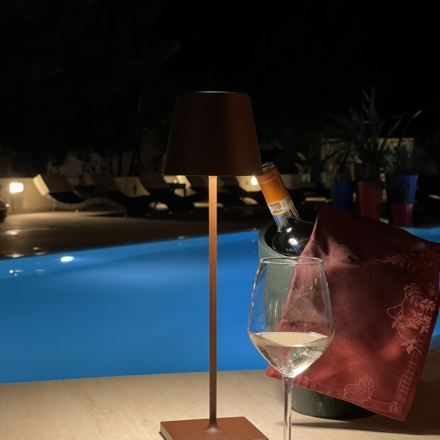 Lampada moderna in rame davanti a una piscina notturna, con bicchiere di vino, bottiglia e tovagliolo rosso.