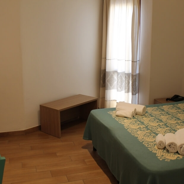 Camera serena con un letto matrimoniale, lenzuolo verde decorato, asciugamani arrotolati e luce naturale che filtra.