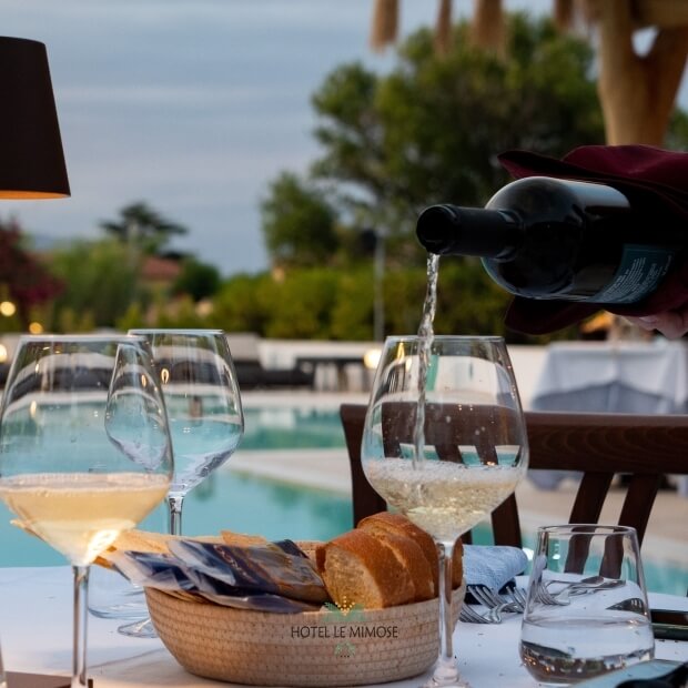 Tavolo da cena con vino bianco, bicchieri, pane e acqua, illuminato da luci, con piscina sfocata sullo sfondo.