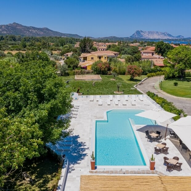 Paesaggio sereno con piscina, alberi e case, monti sullo sfondo e cielo blu, ideale per relax e svago.