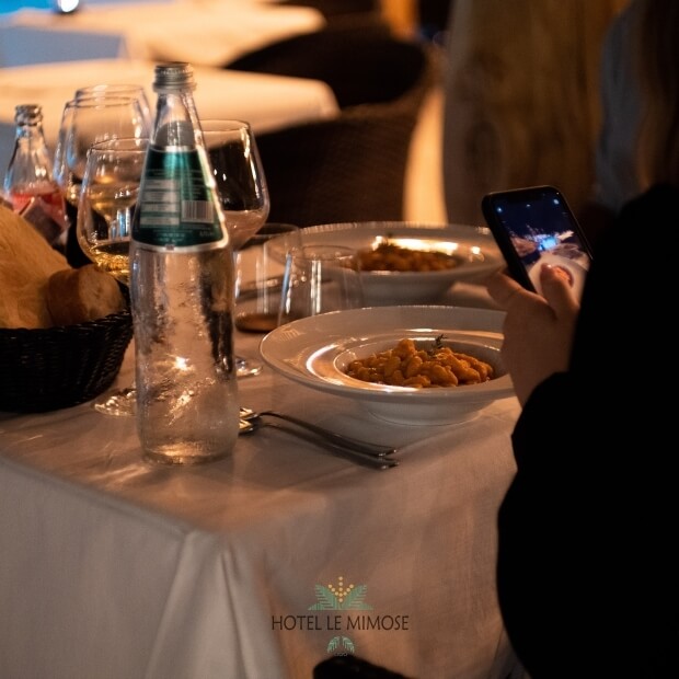 Tavolo da cena con cibo arancione, acqua e pane; persona in maglietta nera scatta foto del piatto con il telefono.