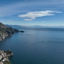 Wide blue ocean under a cloudy sky, with green hills and white buildings on the left.