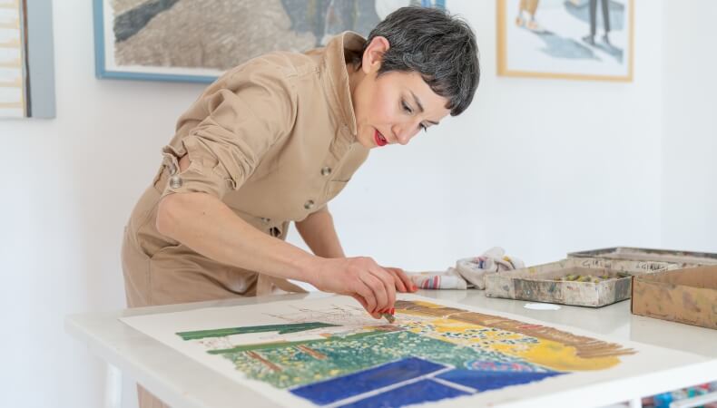 Woman with gray hair painting at a table with art supplies.