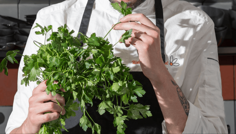Chef in white coat holds fresh parsley, logo visible on coat.