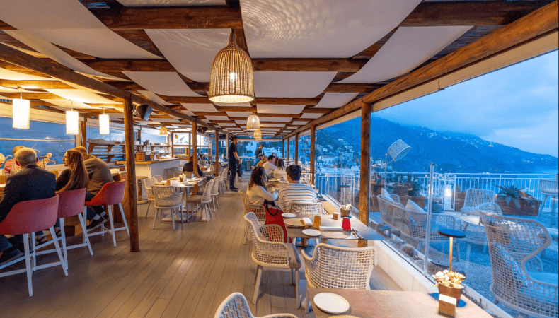 Restaurant patio with diners, wooden structure, coastal town view.