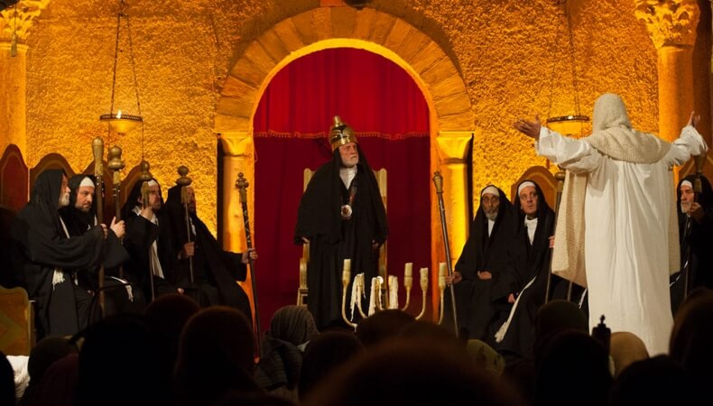 Theatrical scene with a crowned figure on a throne and onlookers.