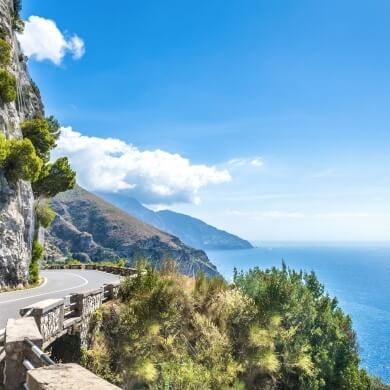 Curved road along a cliff with ocean views and trees.