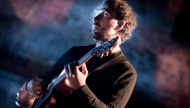 Man with curly hair and beard playing guitar in dim blue and pink lighting.