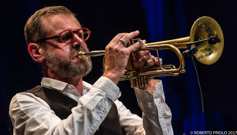 Bearded man in glasses plays trumpet with microphone, blue lighting.