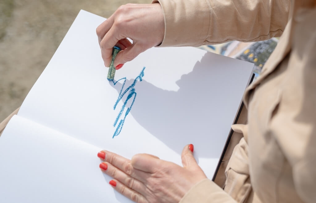 Person with red nails drawing with a blue pastel on a sketchbook.