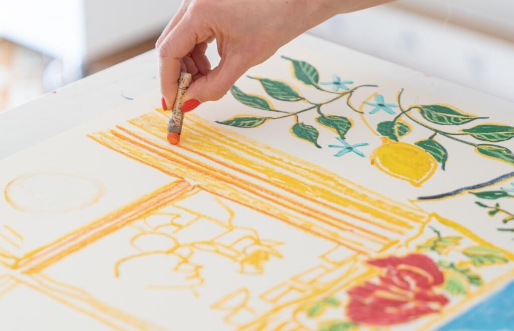 Hand with red nails drawing a lemon and flowers with an orange crayon.