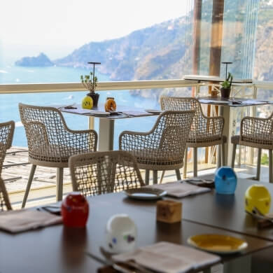 Area pranzo all'aperto con tavoli, vista su acqua e montagne.