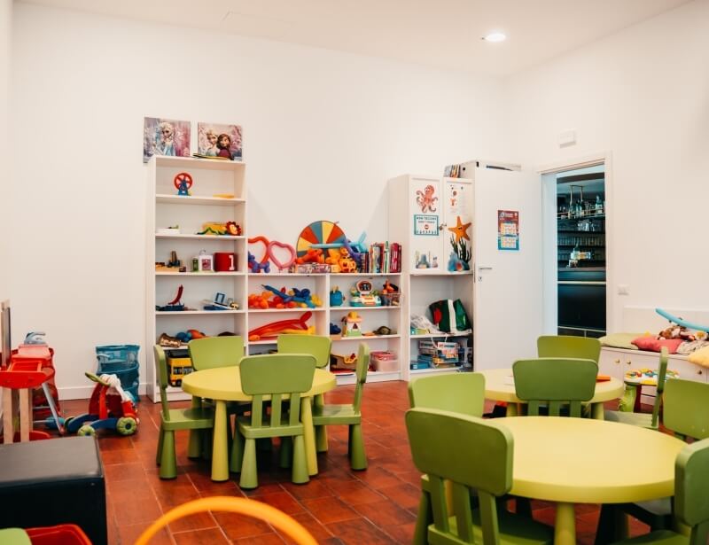 Stanza con scaffali di giocattoli, tavoli gialli e sedie verdi.
