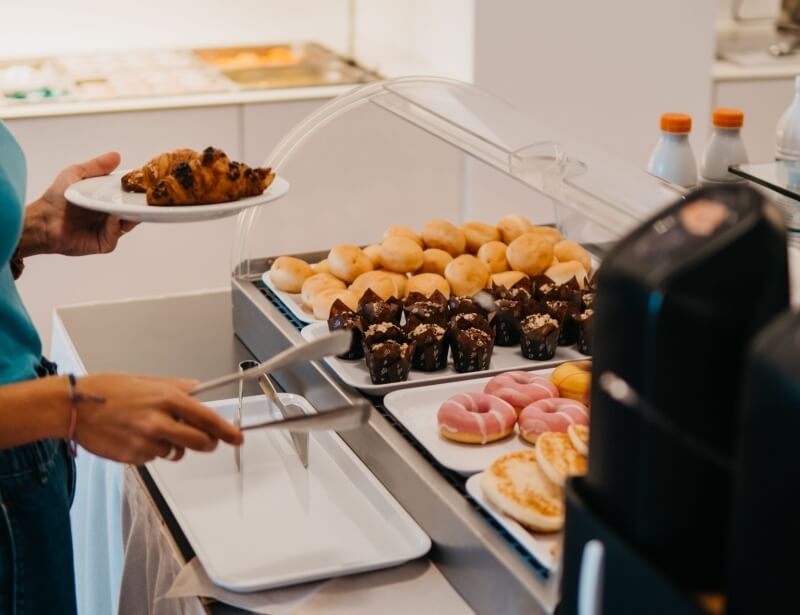 Buffet con croissant, muffin, donuts e pancake; persona con piatto.