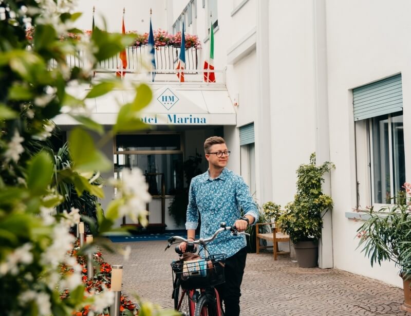 Person walking a red bicycle by "otel Marina" with flags.