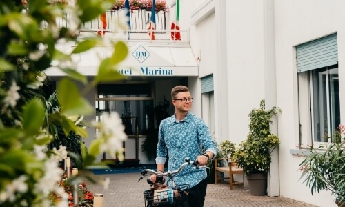Uomo con bicicletta davanti all'Hotel Marina, circondato da piante.