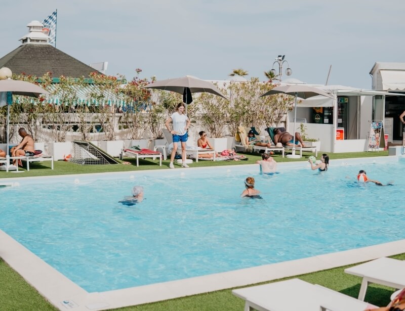 Outdoor pool with swimmers, lounge chairs, and a building nearby.