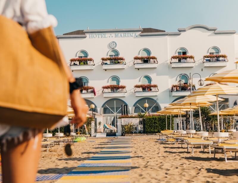 White "HOTEL MARINA" building behind a sandy beach with umbrellas.