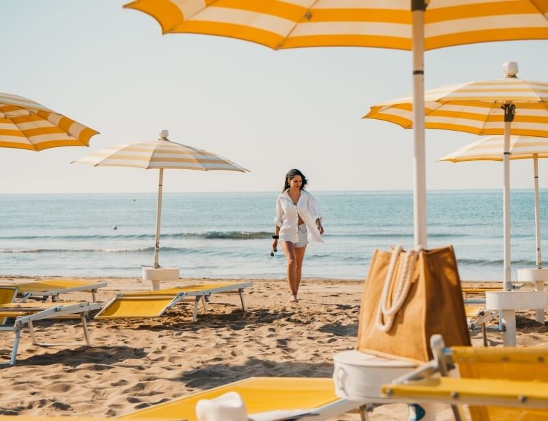 Woman in white shirt and shorts walks on sandy beach with umbrellas.