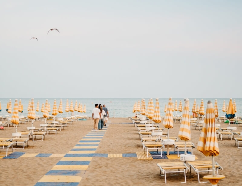 Beach scene with striped umbrellas, lounge chairs, and people walking.