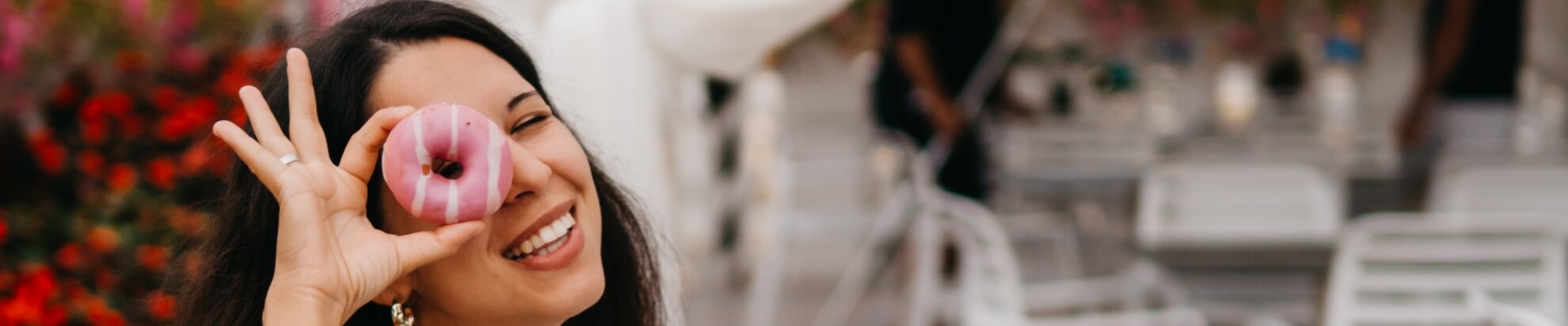 Woman smiling with a donut over her eye, greenery in background.