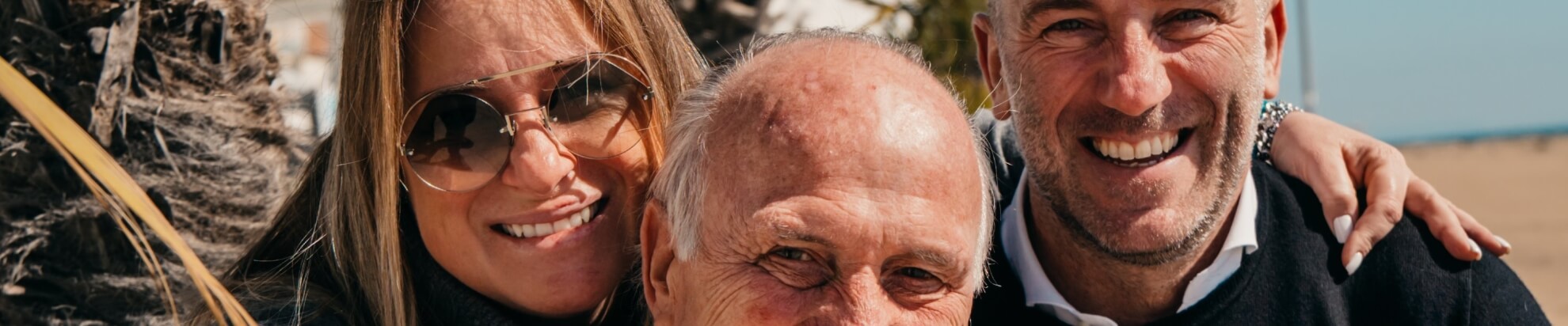 Three people outdoors: a woman in sunglasses, a man in a black sweater, and an older man, with palm fronds behind.