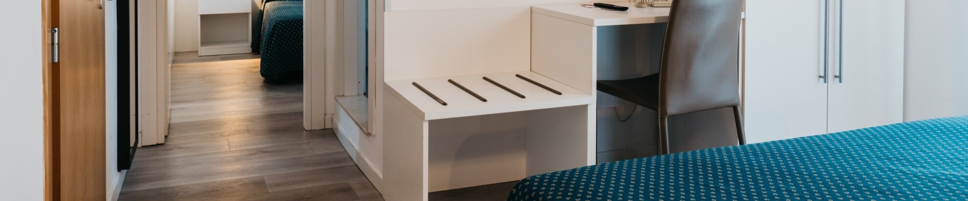 Camera d'hotel con letto teal, scrivania e armadio bianco.