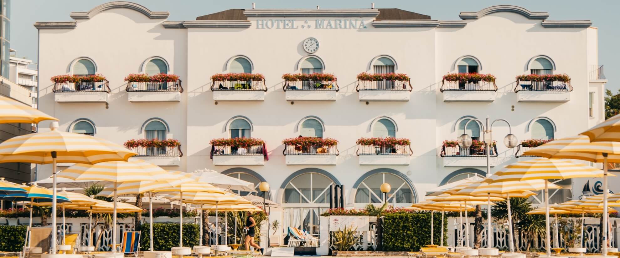 Hotel Marina with white facade, balconies, and striped umbrellas.  
Hotel Marina con facciata bianca, balconi e ombrelloni a strisce.
