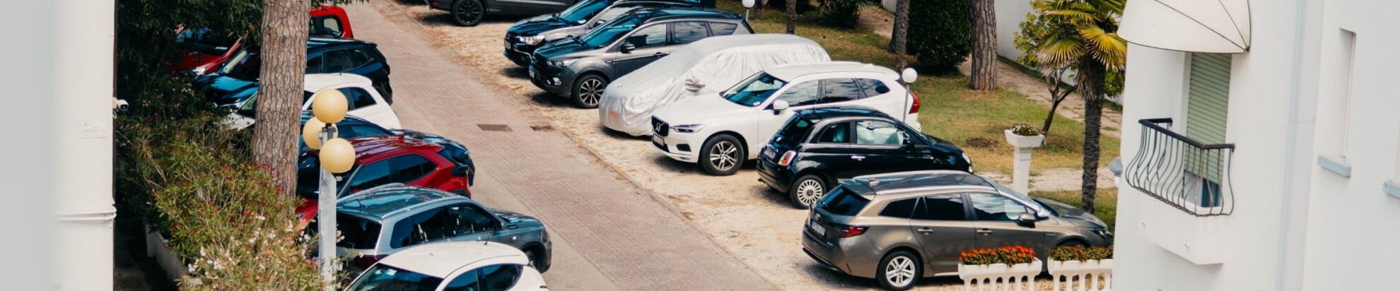 Parking lot with cars under tarps, surrounded by trees and a white building.