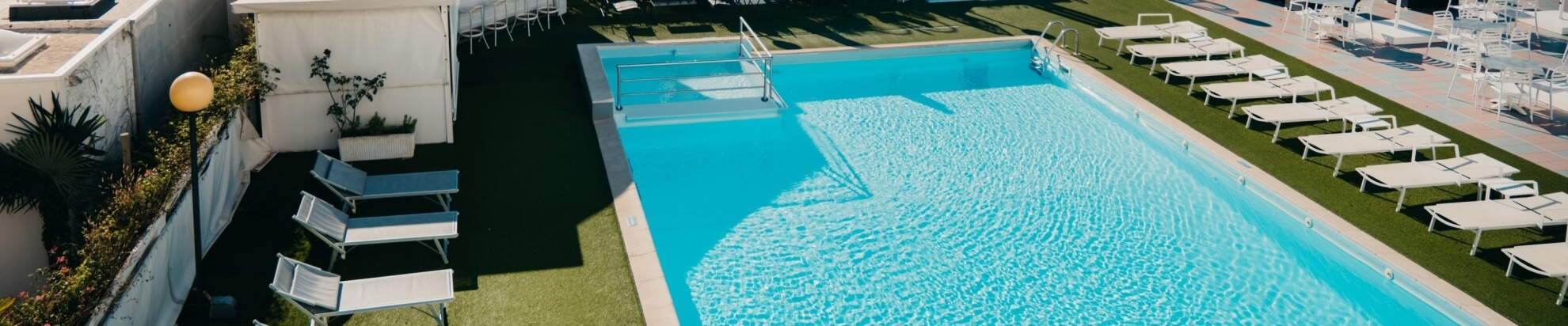 Piscina all'aperto con acqua blu, lettini e verde circostante.