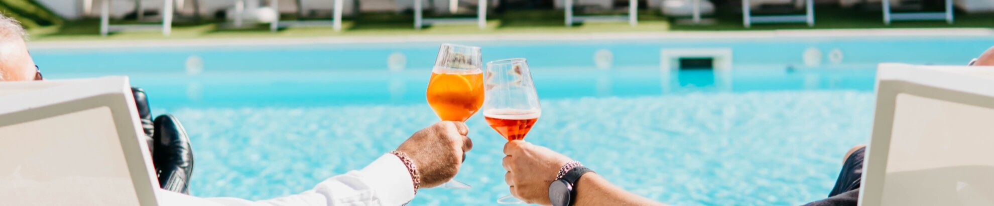 Two hands hold drinks by a pool; one orange, one pinkish-orange.