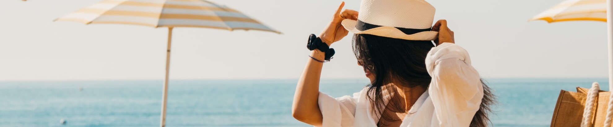Woman in white shirt and hat by the ocean, adjusting her hat.