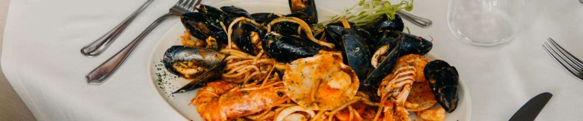 Piatto di spaghetti con frutti di mare e posate su tovaglia bianca.