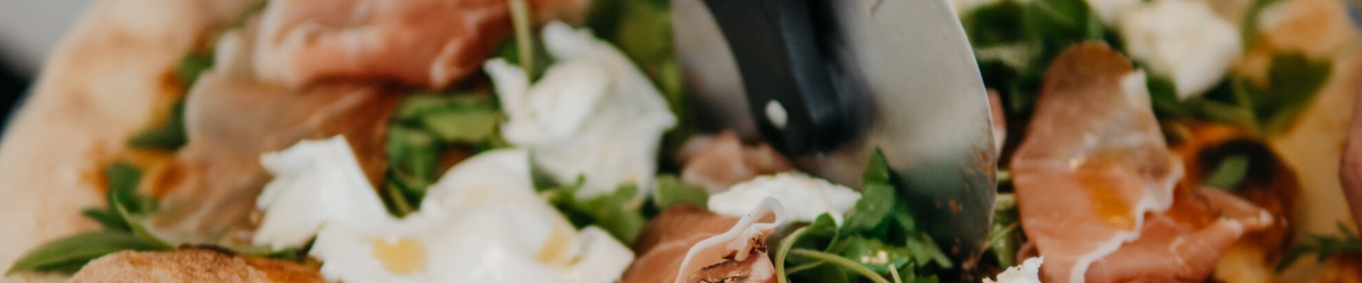 Pizza with prosciutto, cheese, and greens being sliced.