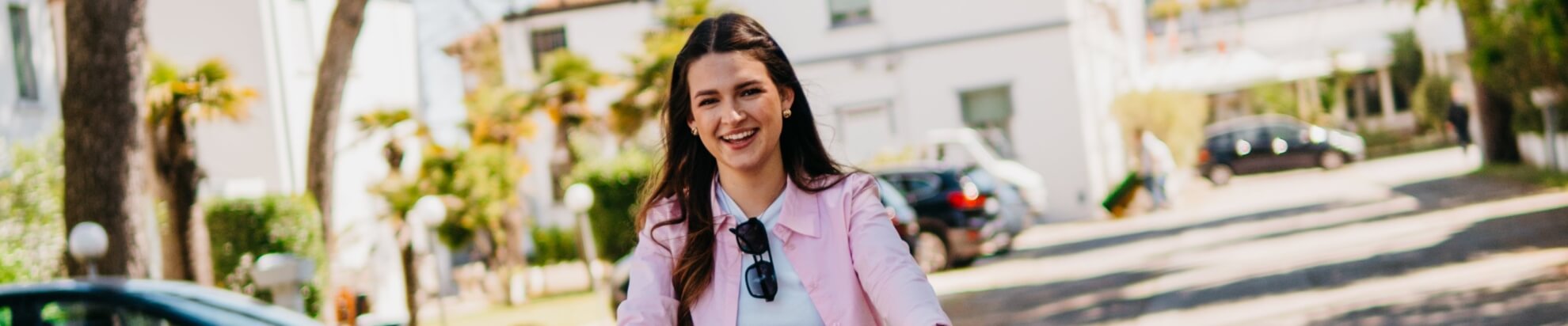 Smiling woman in pink jacket with sunglasses, urban background.
