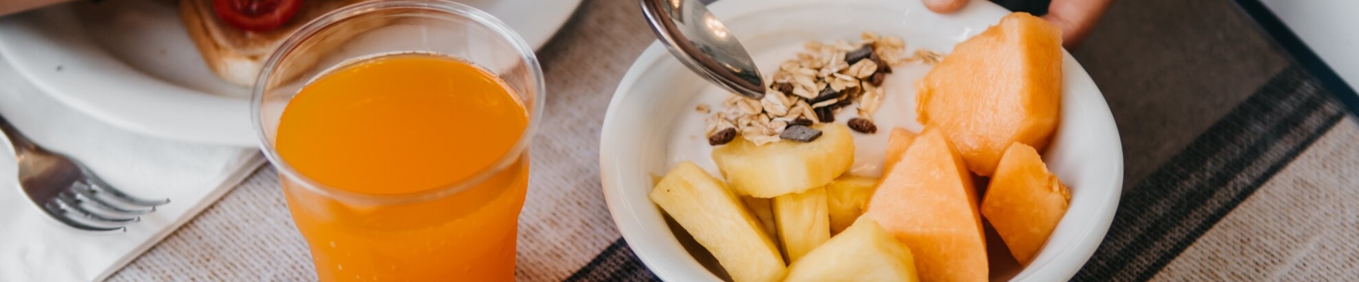 Glass of orange juice beside a bowl of fruit and granola.