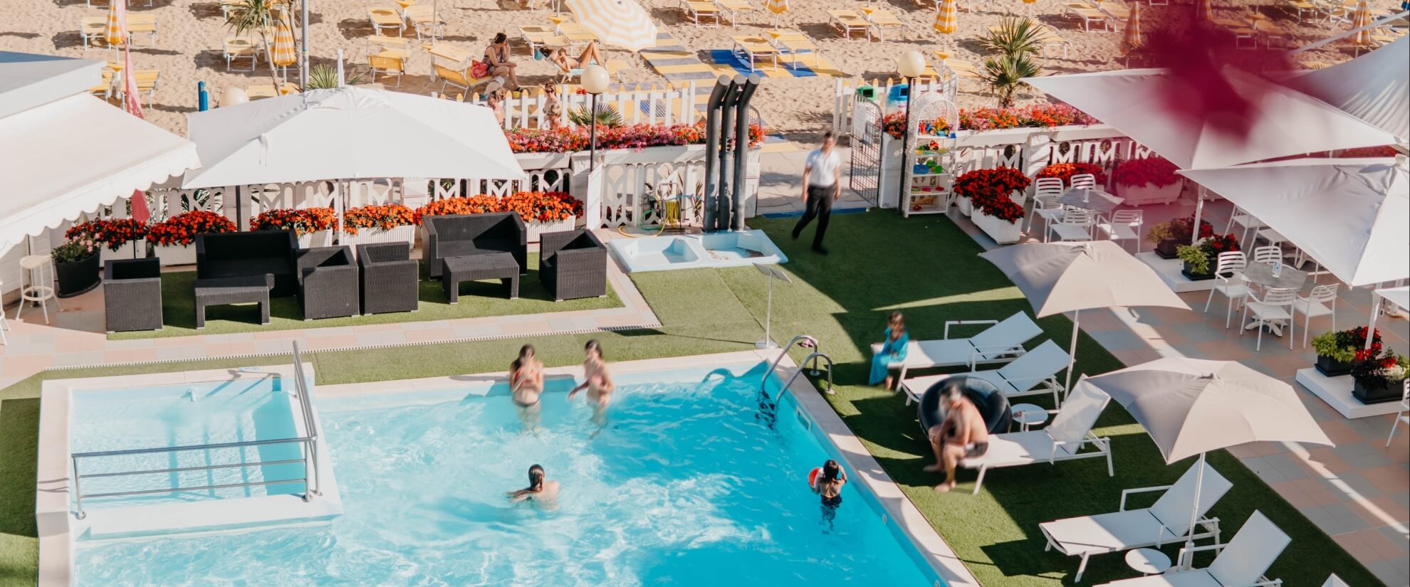 Area piscina all'aperto con persone, lettini e spiaggia sabbiosa.