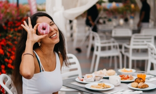 Woman holds a pink donut over her eye, surrounded by breakfast items outdoors.