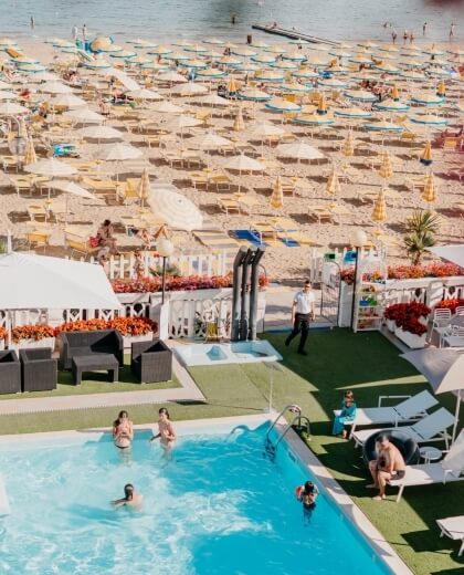 Piscina affollata con lettini, spiaggia sabbiosa e mare sullo sfondo.