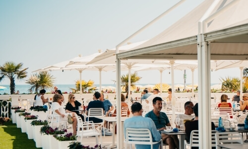 People dining under umbrellas with palm trees and ocean backdrop.