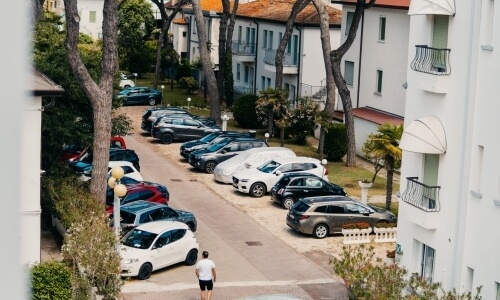 Parking lot with various cars, buildings, trees, and a man walking.